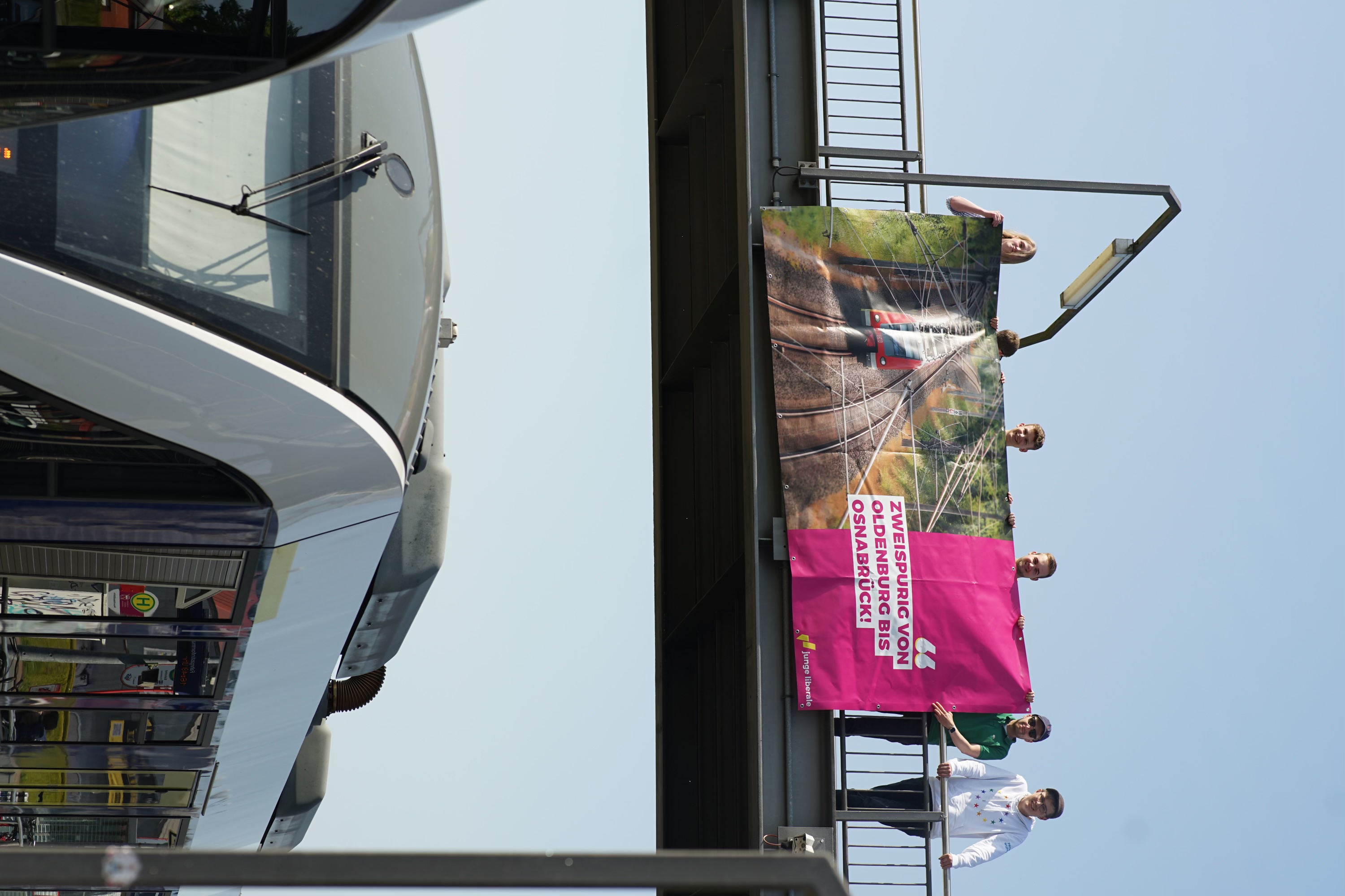 Die Jungliberalen halten das Plakat für den zweispurigen, vollelektrifizierten Ausbau am Bahnhof in Bramsche auf einer Brücke über einen vorbeifahrenden Zug
