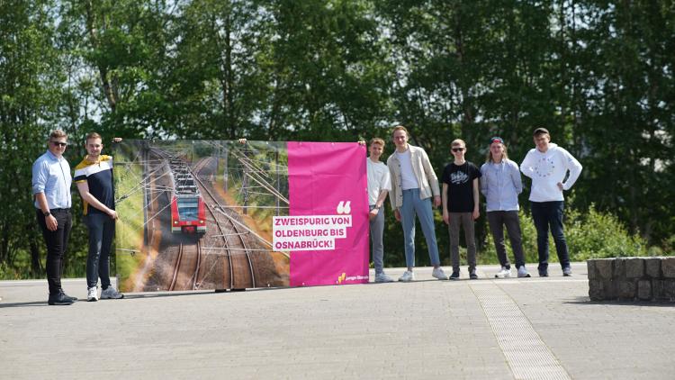 Die Jungliberalen halten das Plakat für den zweispurigen, vollelektrifizierten Ausbau am Bahnhof in Cloppenburg hoch